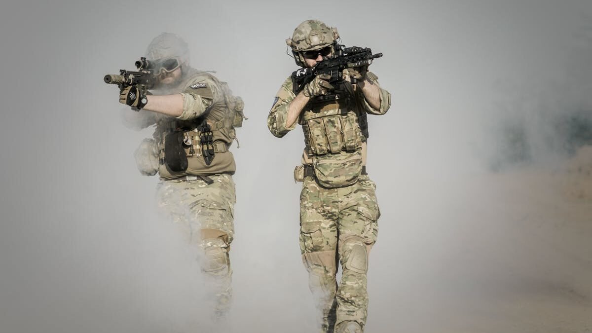 Du kariai su kamufliažinėmis uniformomis ir ginklais juda per dūmais padengtą lauką