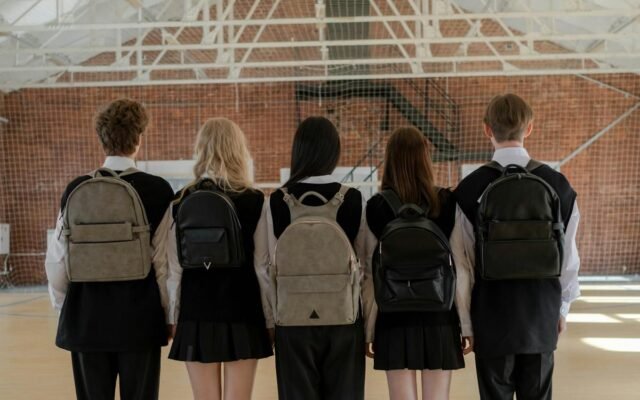 Penkių uniformuotų studentų grupė su kuprinėmis stovi patalpoje nusisukę